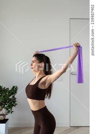 Vertical portrait of a fit young woman performing stretching and fitness exercises with resistance band at home. Female training indoors in sportswear during wellness and healthy lifestyle routine Vertical portrait of a fit young woman performing stretching and fitness exercises with resistance band at home. Female training indoors in sportswear during wellness and healthy lifestyle routine 139987402