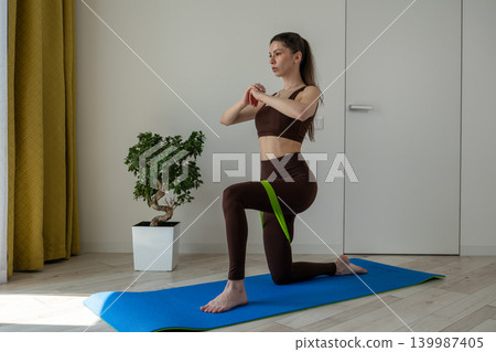 Young woman practicing stretching exercises with fitness resistance band at home. Female doing flexibility and mobility workout in cozy apartment interior. Healthy lifestyle, home fitness, wellness Young woman practicing stretching exercises with fitness resistance band at home. Female doing flexibility and mobility workout in cozy apartment interior. Healthy lifestyle, home fitness, wellness 139987405