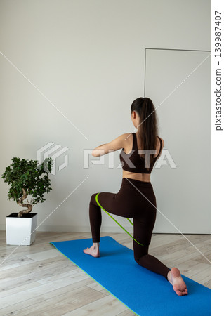 Vertical portrait of a fit young woman performing stretching and fitness exercises with resistance band at home. Female training indoors in sportswear during wellness and healthy lifestyle routine 139987407