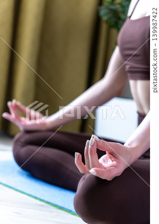 Young woman meditating and practicing yoga at home in a cozy living room. Calm female sitting on yoga mat in lotus pose during morning wellness routine. Concept of mindfulness, healthy lifestyle Young woman meditating and practicing yoga at home in a cozy living room. Calm female sitting on yoga mat in lotus pose during morning wellness routine. Concept of mindfulness, healthy lifestyle 139987422