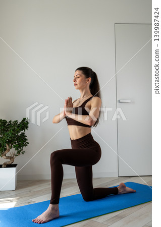 Vertical portrait of a peaceful young woman meditating and practicing yoga indoors. Female sitting cross legged on a yoga mat in a cozy apartment during morning wellness routine. Concept of Vertical portrait of a peaceful young woman meditating and practicing yoga indoors. Female sitting cross legged on a yoga mat in a cozy apartment during morning wellness routine. Concept of 139987424