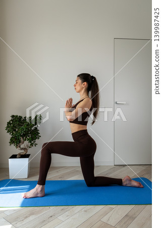 Vertical portrait of a peaceful young woman meditating and practicing yoga indoors. Female sitting cross legged on a yoga mat in a cozy apartment during morning wellness routine. Concept of Vertical portrait of a peaceful young woman meditating and practicing yoga indoors. Female sitting cross legged on a yoga mat in a cozy apartment during morning wellness routine. Concept of 139987425