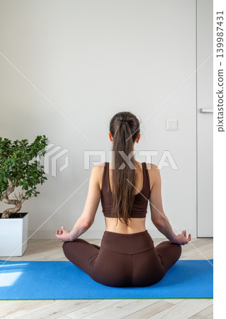 Vertical portrait of a peaceful young woman meditating and practicing yoga indoors. Female sitting cross legged on a yoga mat in a cozy apartment during morning wellness routine. Concept of 139987431