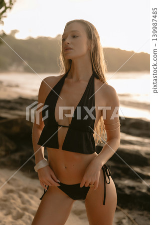 Blonde woman in black bikini posing on rocky beach at sunset, sensual portrait 139987485