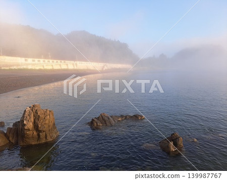 清晨的薄霧籠罩著岩手縣宮古市三陸海岸藤之川海灘的沙灘和防波堤,沐浴在冉冉升起的太陽的金色光芒中。 清晨的薄霧籠罩著岩手縣宮古市三陸海岸藤之川海灘的沙灘和防波堤,沐浴在冉冉升起的太陽的金色光芒中。 139987767