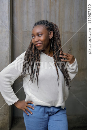 Casual portrait of a smiling young Black woman with long braids looking away against a concrete wall Casual portrait of a smiling young Black woman with long braids looking away against a concrete wall 139987860