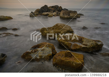Rocky shoreline at dusk with gentle waves kissing the coast and a serene sky Rocky shoreline at dusk with gentle waves kissing the coast and a serene sky 139988218