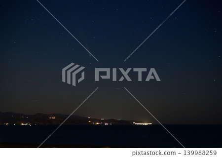 Starry night sky over the serene coastline with distant lights twinkling along the shore and a comet trailing through the cosmos Starry night sky over the serene coastline with distant lights twinkling along the shore and a comet trailing through the cosmos 139988259