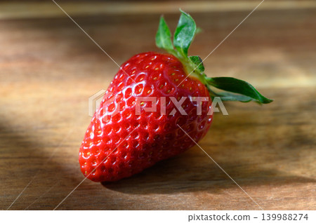 Freshly harvested strawberry resting on a rustic wooden surface under soft sunlight, inviting senses with its vibrant color and texture Freshly harvested strawberry resting on a rustic wooden surface under soft sunlight, inviting senses with its vibrant color and texture 139988274