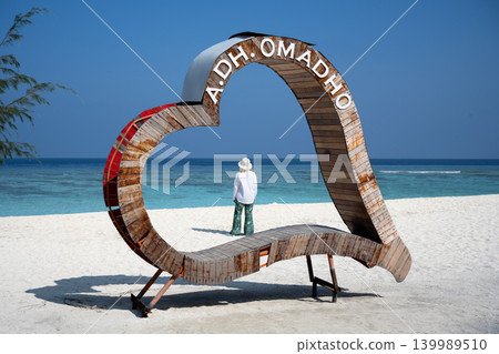Woman standing inside a large wooden heart structure on a white sand beach in Omadhoo Maldives. Tourist looking at the turquoise ocean. Summer vacation travel concept Woman standing inside a large wooden heart structure on a white sand beach in Omadhoo Maldives. Tourist looking at the turquoise ocean. Summer vacation travel concept 139989510