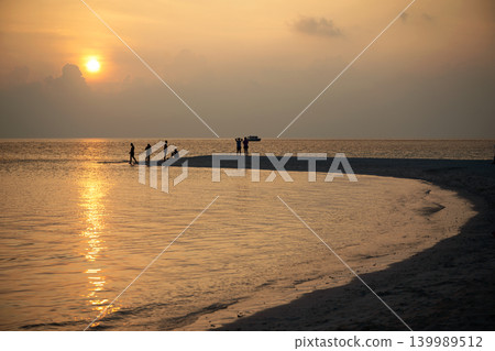 Silhouetted people on a sandbar at sunset in Omadhoo Maldives. Group of tourists enjoying the golden hour on a sea tropical beach. Vacation, ocean and travel concept. Silhouetted people on a sandbar at sunset in Omadhoo Maldives. Group of tourists enjoying the golden hour on a sea tropical beach. Vacation, ocean and travel concept. 139989512