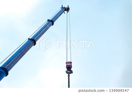 Blue crane arm, boom with hanging hook against clear sky. Construction and lifting heavy building materials, large components, and machinery Blue crane arm, boom with hanging hook against clear sky. Construction and lifting heavy building materials, large components, and machinery 139989547
