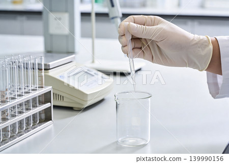 Scientist carefully adds liquid drop to beaker using pipette in a sterile laboratory setting Scientist carefully adds liquid drop to beaker using pipette in a sterile laboratory setting 139990156
