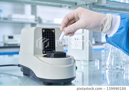 Scientist in lab coat and gloves places sample tube into laboratory heating device 139990180