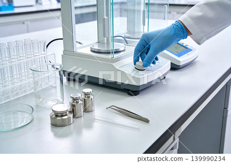 Scientist's gloved hand calibrating precise laboratory scale with weights and glassware Scientist's gloved hand calibrating precise laboratory scale with weights and glassware 139990234