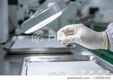 Scientist inspects small white cap under bright magnifying lamp in laboratory setting Scientist inspects small white cap under bright magnifying lamp in laboratory setting 139990369