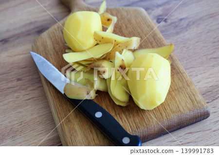 Two peeled potatoes with their brown skins and a knife on a wooden cutting board. Vegetables are prepared for cooking a healthy family dinner Two peeled potatoes with their brown skins and a knife on a wooden cutting board. Vegetables are prepared for cooking a healthy family dinner 139991783