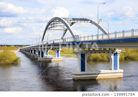 Arched metal road bridge across the river, connecting the two banks. Modern concept for road infrastructure, logistics, and transportation. Arched metal road bridge across the river, connecting the two banks. Modern concept for road infrastructure, logistics, and transportation. 139991784