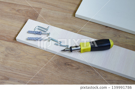 Screwdriver with a black and yellow plastic handle, plastic dowels, screws, and furniture boards on the floor, ready for furniture assembly. Screwdriver with a black and yellow plastic handle, plastic dowels, screws, and furniture boards on the floor, ready for furniture assembly. 139991786