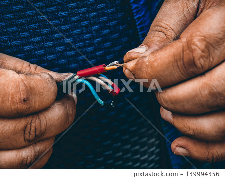 Worker hands carefully holding and manipulating stripped electrical wires, demonstrating the maintenance process of electrical connections for repair and setup. 139994356