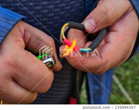 Person's hands using a gas lighter to apply heat to red heat shrink tubing, securing and insulating a spliced electrical wire connection, ensuring a safe and durable repair outdoors. Person's hands using a gas lighter to apply heat to red heat shrink tubing, securing and insulating a spliced electrical wire connection, ensuring a safe and durable repair outdoors. 139994367