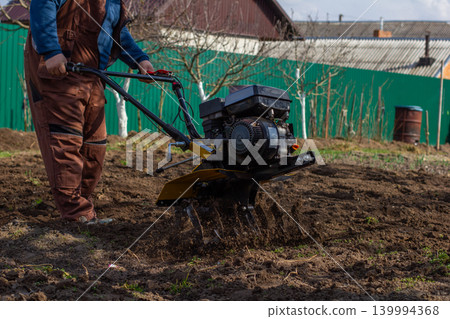 Man in overalls operating a walk-behind tractor to till rich brown loam in a spring vegetable garden, cultivating soil and preparing seedbed for planting and growth. Man in overalls operating a walk-behind tractor to till rich brown loam in a spring vegetable garden, cultivating soil and preparing seedbed for planting and growth. 139994368