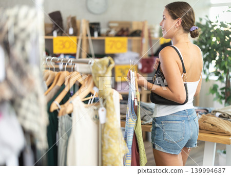 Positive young woman choosing something for summer in clothing store Positive young woman choosing something for summer in clothing store 139994687