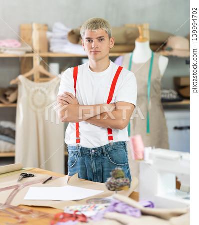 Stylish young male dressmaker posing near work place with sewing machine and tools, welcoming clients in own sewing workshop Stylish young male dressmaker posing near work place with sewing machine and tools, welcoming clients in own sewing workshop 139994702