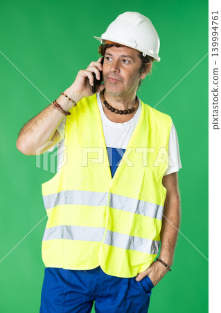 engineer wearing a safety helmet and a reflective vest talking on a mobile phone on a green background engineer wearing a safety helmet and a reflective vest talking on a mobile phone on a green background 139994761