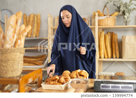 Girl in traditional Muslim clothing selects and places fresh hot croissants in plastic bag Girl in traditional Muslim clothing selects and places fresh hot croissants in plastic bag 139994774