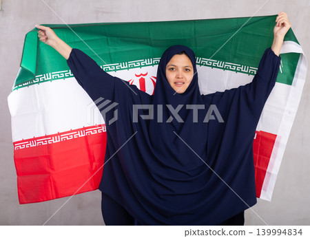 Young Muslim woman in blue hijab holds unfurled flag of Iran. Young Muslim woman in blue hijab holds unfurled flag of Iran. 139994834