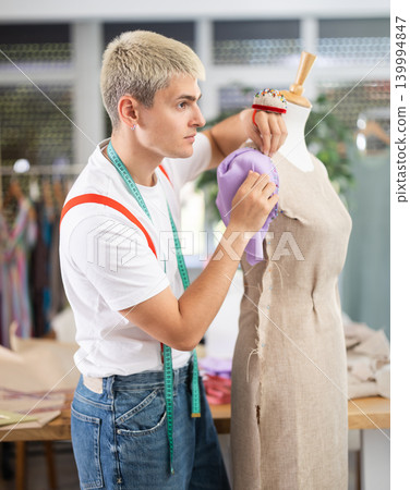 Experienced young male fashion designer working with fabric and mannequin while sewing fashionable dress in sewing workshop 139994847