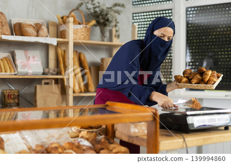 Middle-aged Muslim saleswoman weights croissants in bakery Middle-aged Muslim saleswoman weights croissants in bakery 139994860
