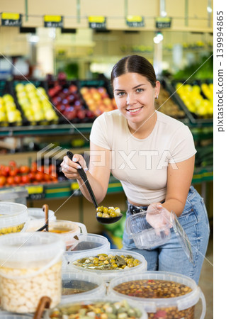 Positive woman putting pickled olives into a container in the grocery section of supermarket 139994865