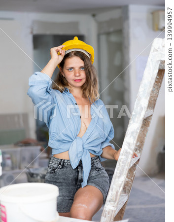 Seductive woman in hardhat and denim shirt posing next to a stepladder in room being renovated Seductive woman in hardhat and denim shirt posing next to a stepladder in room being renovated 139994957