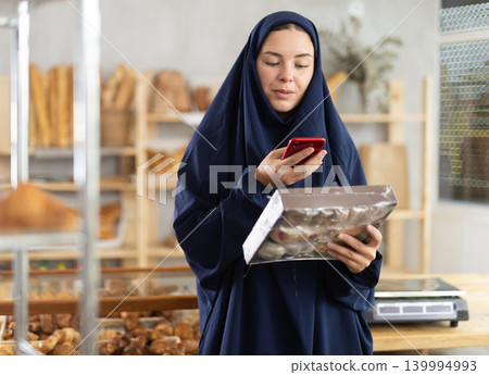 European Muslim woman in hijab customer at bakery scans QR code on cookie package using phone camera European Muslim woman in hijab customer at bakery scans QR code on cookie package using phone camera 139994993