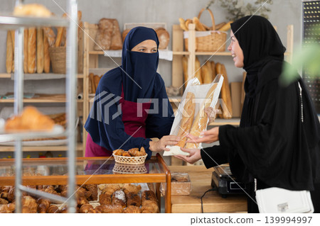 Female bakery salesperson in traditional Islamic dress serves bag of baguettes and bread to a girl in niqab Female bakery salesperson in traditional Islamic dress serves bag of baguettes and bread to a girl in niqab 139994997