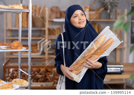 Asian Muslim woman buyer with baguettes in plastic package stands in trading floor of bakery Asian Muslim woman buyer with baguettes in plastic package stands in trading floor of bakery 139995114
