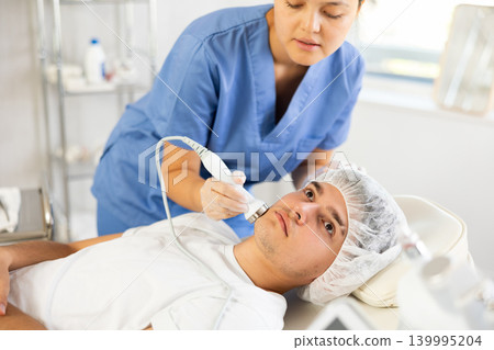 Young woman cosmetologist doing ultrasound procedure to young man Young woman cosmetologist doing ultrasound procedure to young man 139995204
