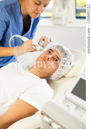 Young woman cosmetologist doing moisturizing procedure to young man Young woman cosmetologist doing moisturizing procedure to young man 139995212