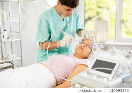 Young male cosmetologist doing exfoliating procedure to young woman Young male cosmetologist doing exfoliating procedure to young woman 139995253