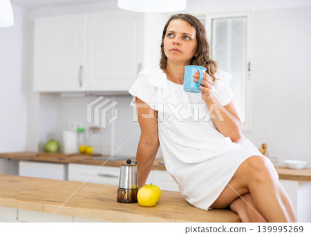 Woman sitting on kitchen countertop, drinking coffee Woman sitting on kitchen countertop, drinking coffee 139995269