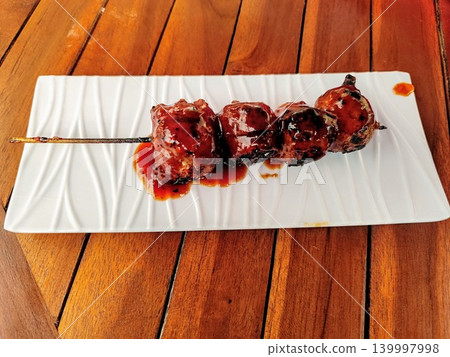 Grilled meatballs on skewers and served with sweet and spicy sauce on a white square plate with a wooden table background Grilled meatballs on skewers and served with sweet and spicy sauce on a white square plate with a wooden table background 139997998