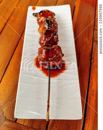 Grilled meatballs on skewers and served with sweet and spicy sauce on a white square plate with a wooden table background Grilled meatballs on skewers and served with sweet and spicy sauce on a white square plate with a wooden table background 139997999