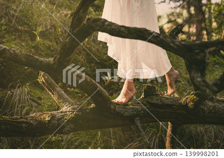Faceless barefoot woman in white vintage dress walking on a mossy fallen tree trunk in enchanted forest, atmospheric cinematic, close-up shot. Faceless barefoot woman in white vintage dress walking on a mossy fallen tree trunk in enchanted forest, atmospheric cinematic, close-up shot. 139998201