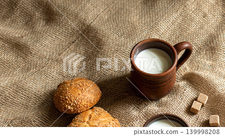 Two golden sesame seed buns, a traditional dark clay mug with fresh milk and brown sugar cubes on a textured burlap, copy space. Two golden sesame seed buns, a traditional dark clay mug with fresh milk and brown sugar cubes on a textured burlap, copy space. 139998208