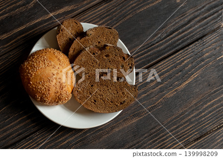 Sesame seed bun and several slices of dark, rustic rye bread on white plate on textured, dark brown wooden table, copy space. Sesame seed bun and several slices of dark, rustic rye bread on white plate on textured, dark brown wooden table, copy space. 139998209