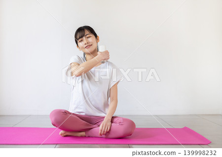 A young Japanese woman self-massages her shoulders with a massage gun on a yoga mat. A young Japanese woman self-massages her shoulders with a massage gun on a yoga mat. 139998232