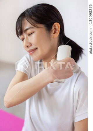一位年輕的日本女性在瑜珈墊上用按摩槍按摩肩膀。 139998236