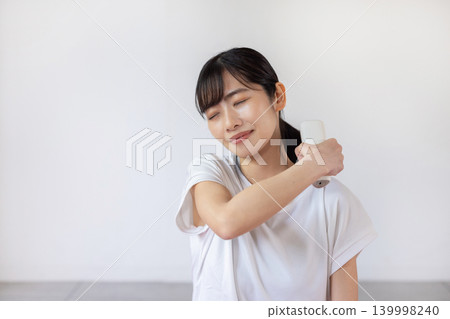 一位年輕的日本女性在瑜珈墊上用按摩槍按摩肩膀。 139998240